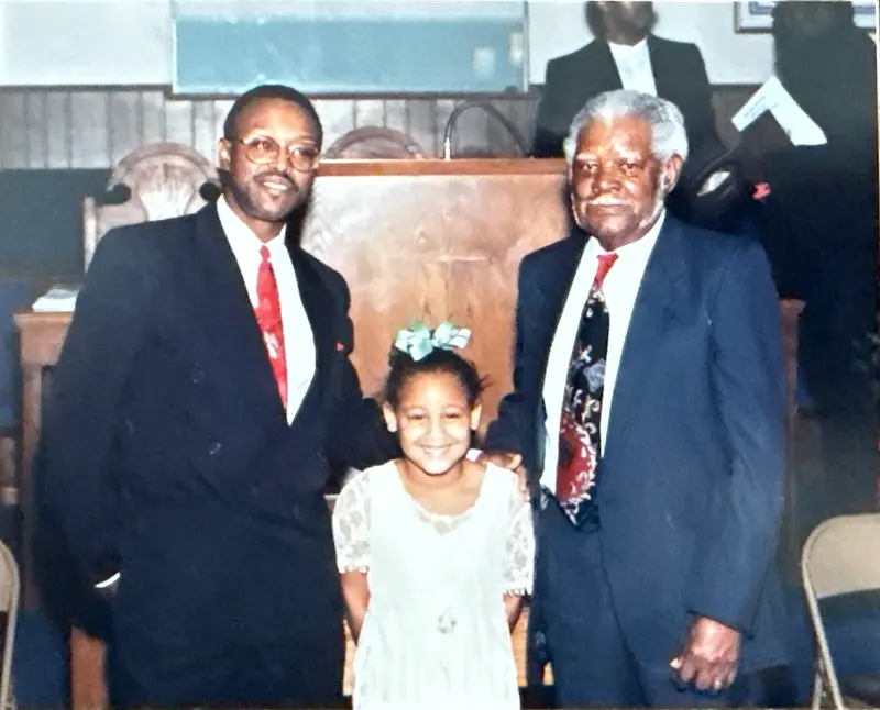 Photo of two men in suits standing with a smiling young girl in front of the pulpit